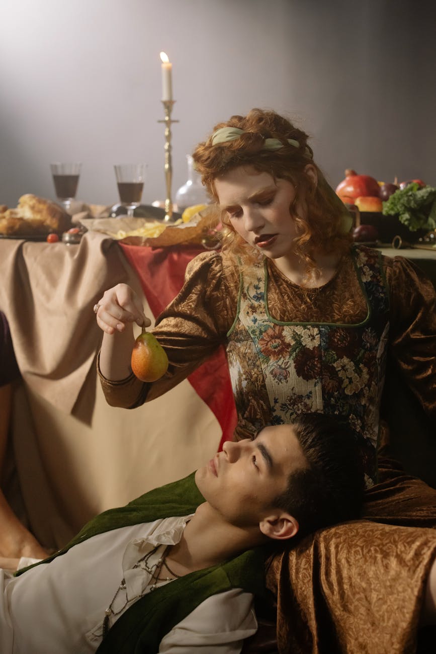 Stock photo of couple in medieval garb lounging on the floor in front of a table bearing a feast with one dangling a piece of fruit by the stem over the other. Pexels.com