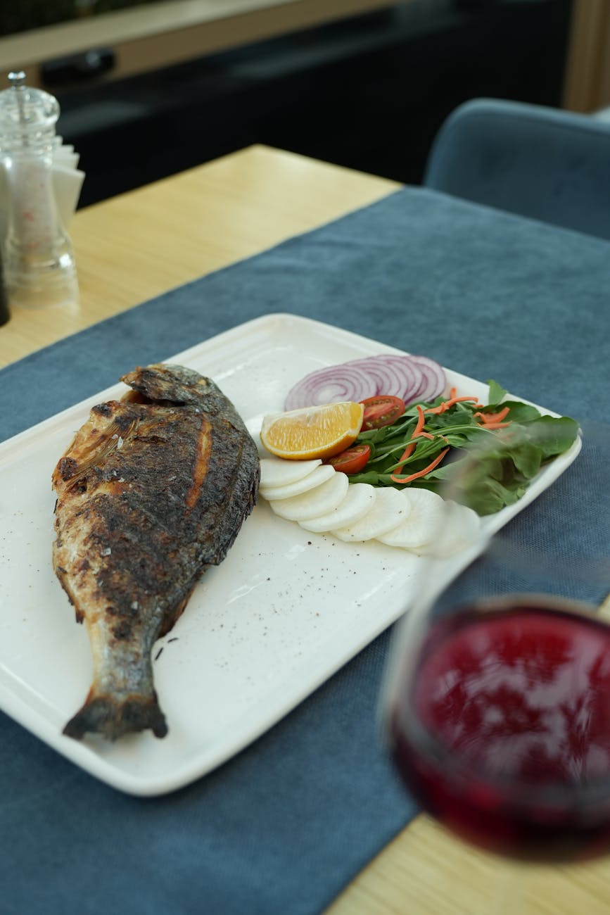 Stock photo of blackened whole fish meal presentation with salad on a plate and a glass of wine from Pexels.com