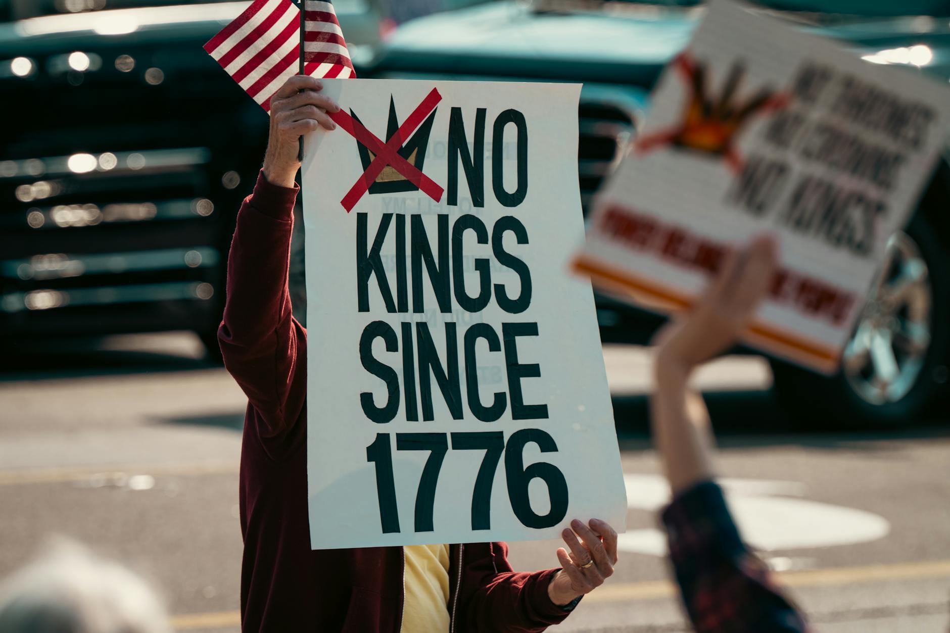Stock photo of a person holding a sign that reads No kings since 1776 and a U.S. flag from Pexels.com