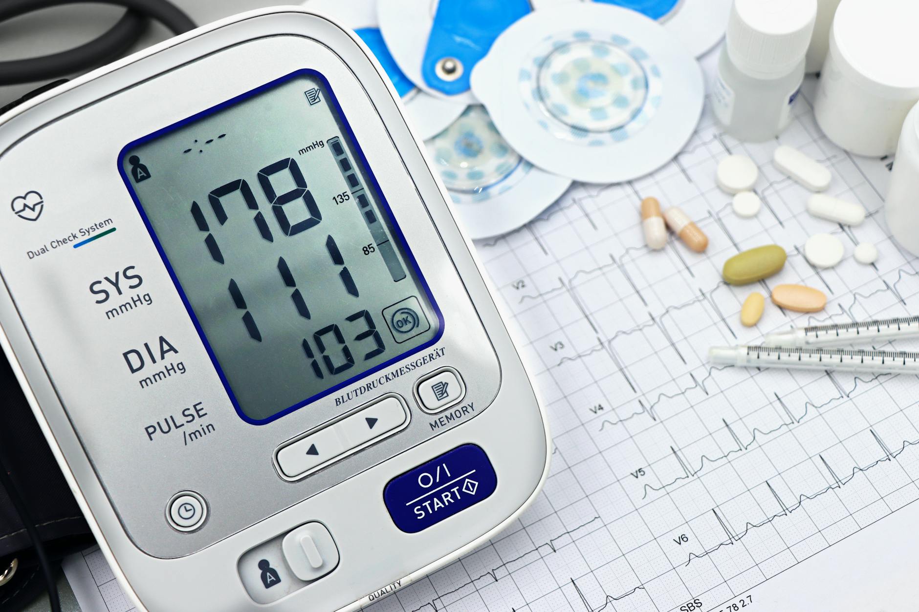 Stock photo of a digital blood pressure monitor with a reading of 178 over 111 with a heart rate of 103 on a table with printouts, pills, syringes, pads, and bottles from Pexels