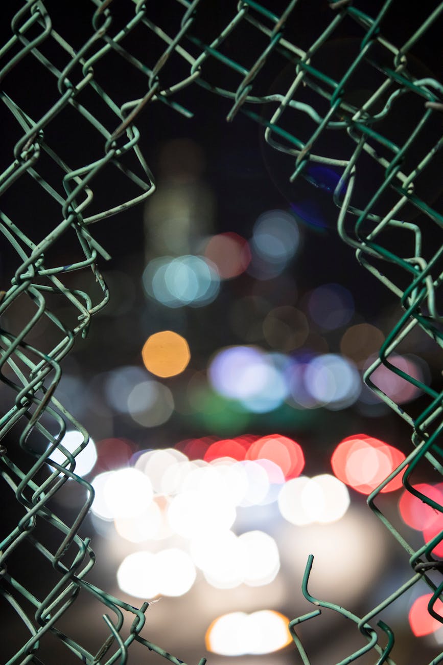 Stock photo of blurred lights through a hole cut in a chain link fence from Pexels.com