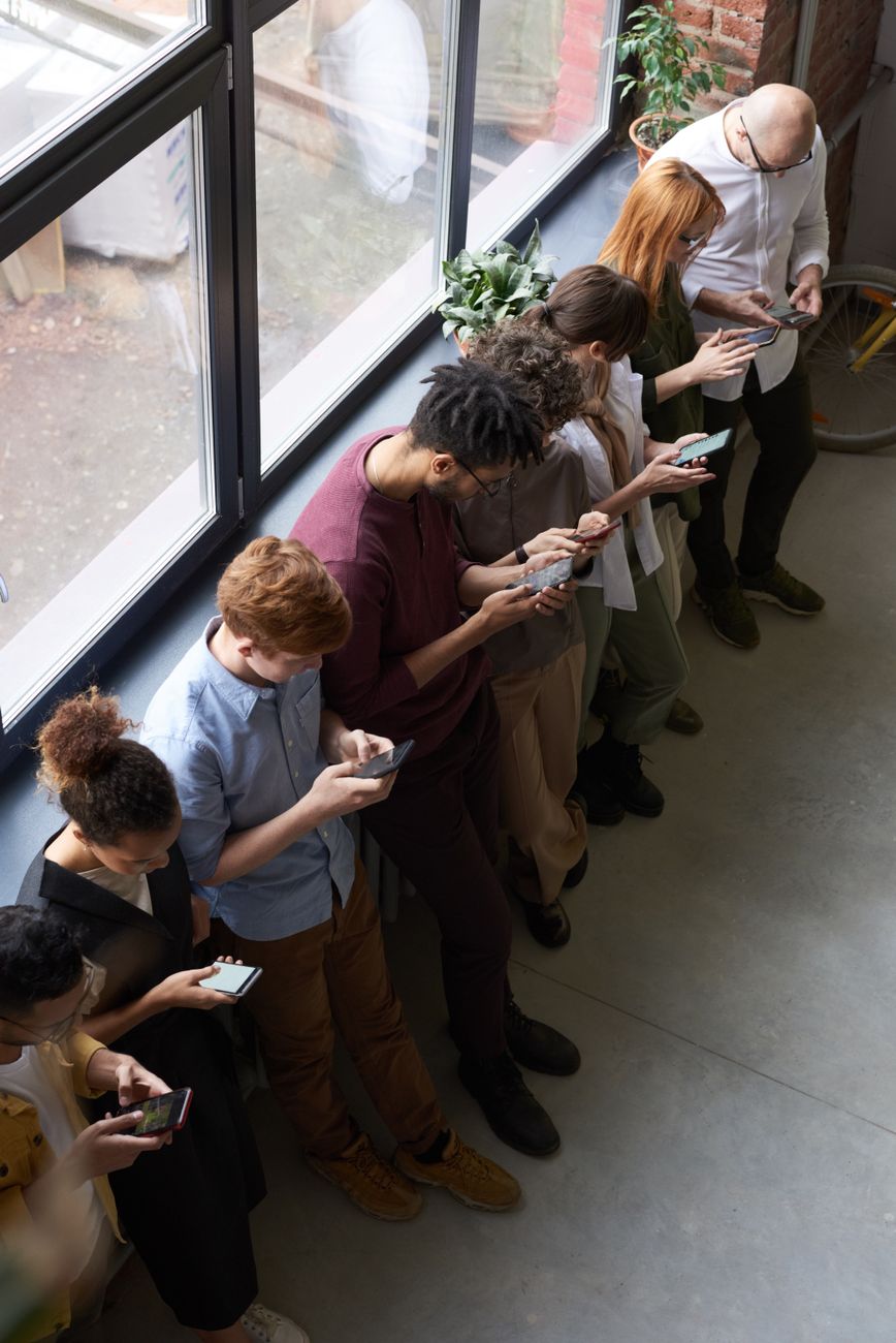 Stock photo of a  line of young people looking at their phones while turning their back to a large window to the world from Pexels.com 