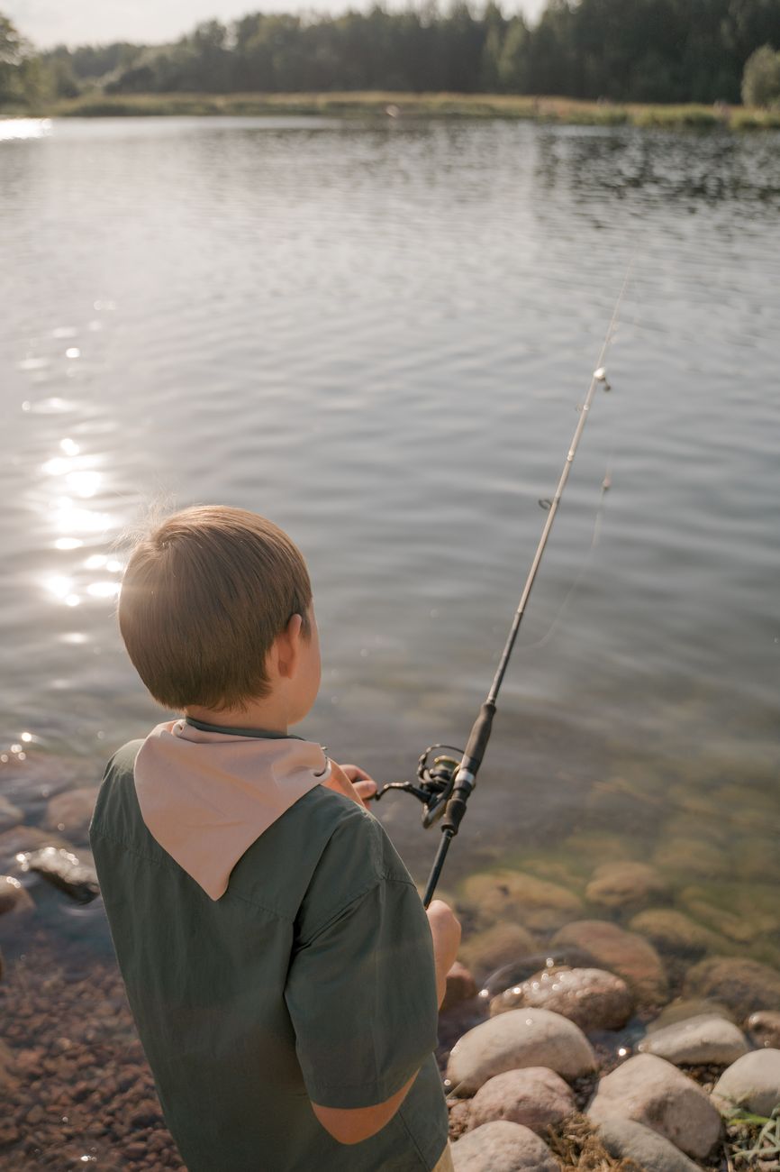 Stock photo of a young person holding a fishing pole on the rocky shore of a lake from Pexels.com