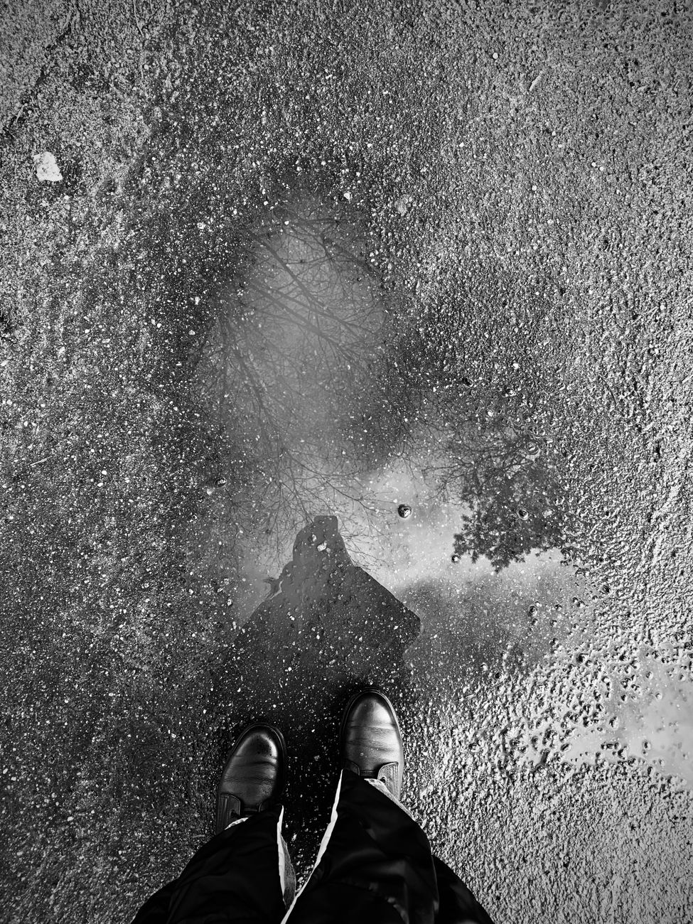 Stock black-and-white street photograph captures a first-person perspective of a person standing on wet asphalt with their shoes positioned near a puddle reflecting a shadow silhouette from Pexels.com