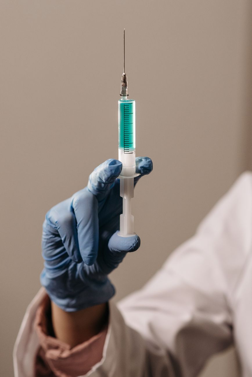 Stock photo of a person wearing blue medical gloves holding a plastic syringe filled with a green liquid from Pexels.com 
