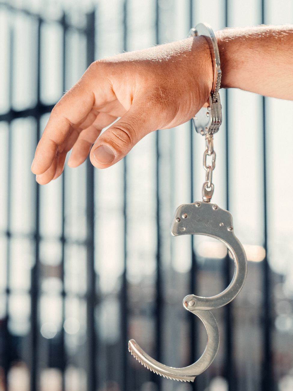  Stock photo of metallic steel handcuffs hanging open from a person's wrist against a blurred prison bar background from Pexels.com
