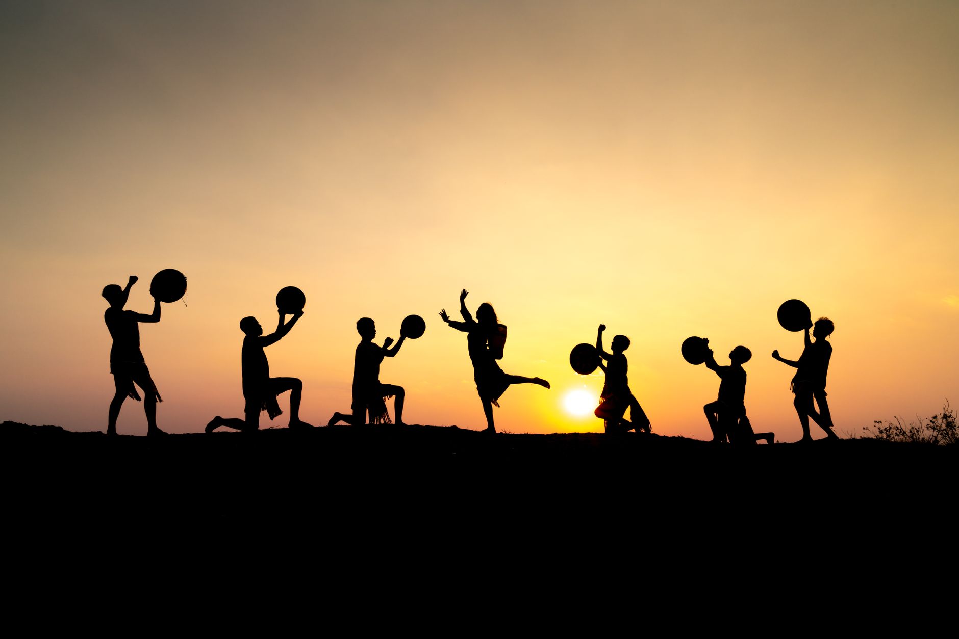 Stock photo shows a group of people silhouetted against a bright sunset while performing a traditional cultural dance with instruments from Pexel.com