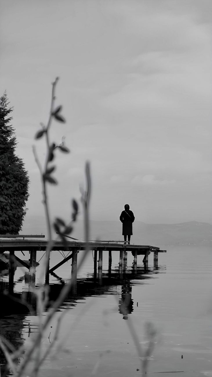 Stock black-and-white photo of a  contemplative scene with a figure standing at the end of a wooden pier or dock extending into a calm body of water, with out-of-focus branches in the foreground from Pexels.com