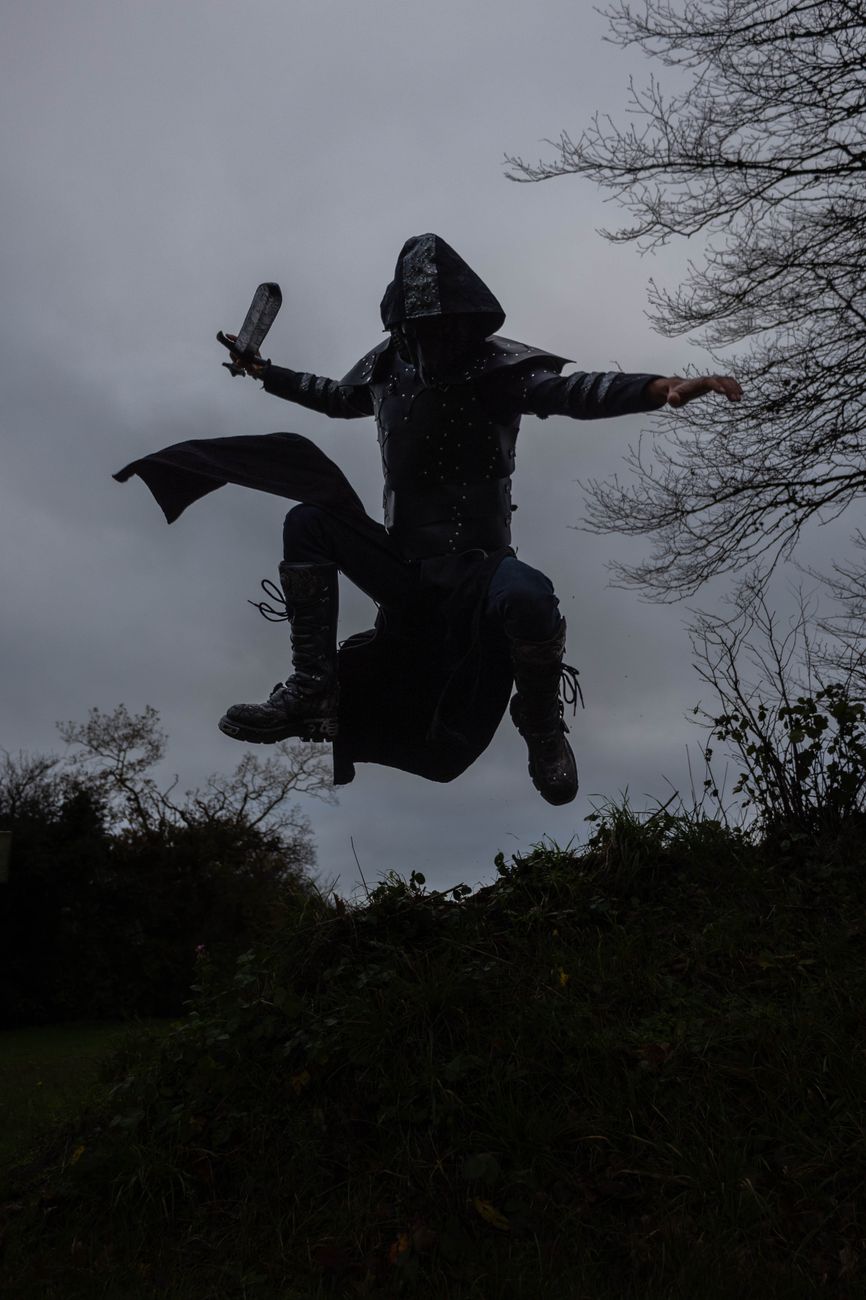 Stock photo of a person dressed in dark tactical or fantasy-style gear with a hood, captured mid-air leaping above a bushy landscape under an overcast sky from Pexels.com