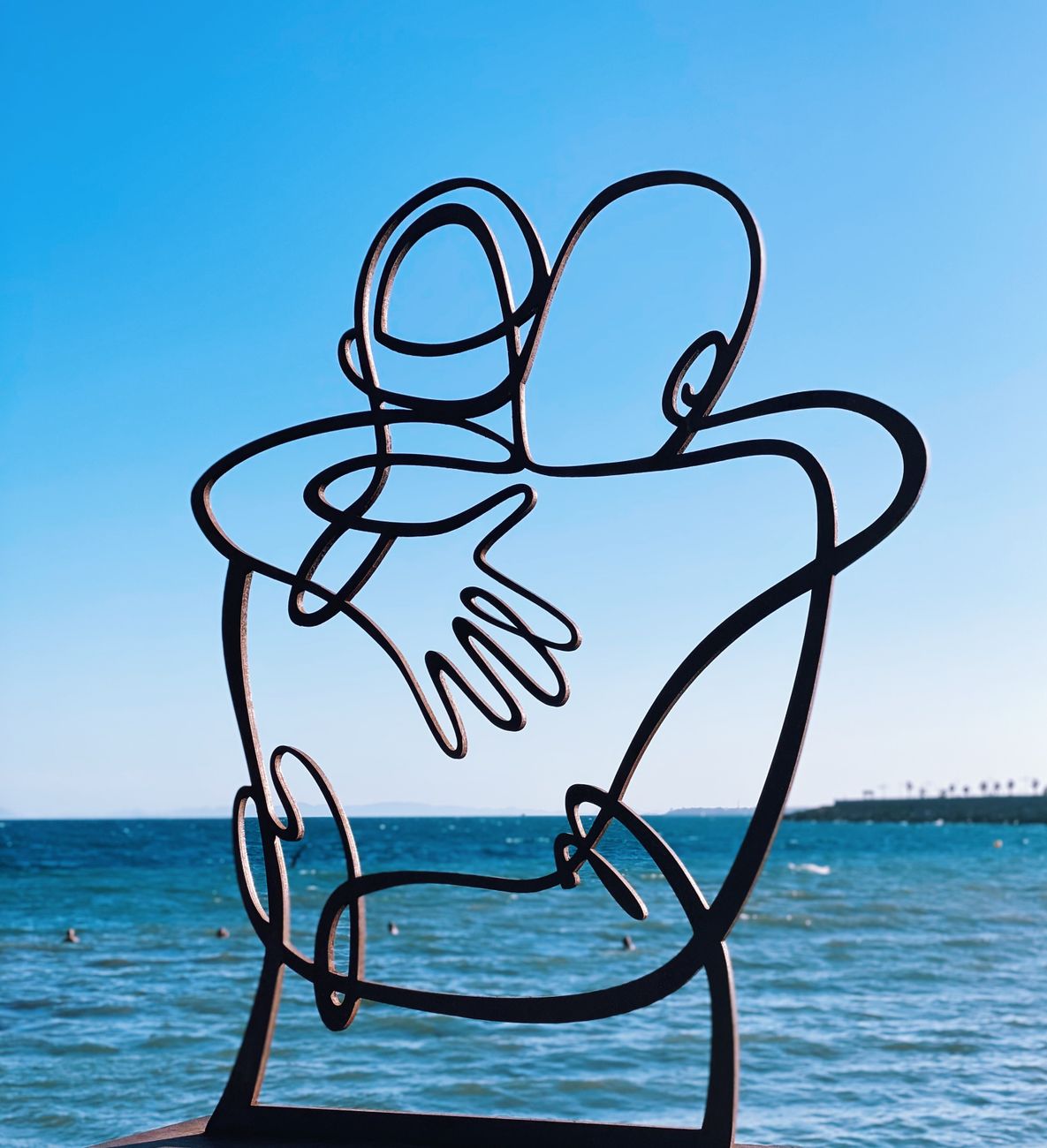 Stock photo shows a metal line-art sculpture of two figures embracing against a backdrop of the sea from Pexels.com 