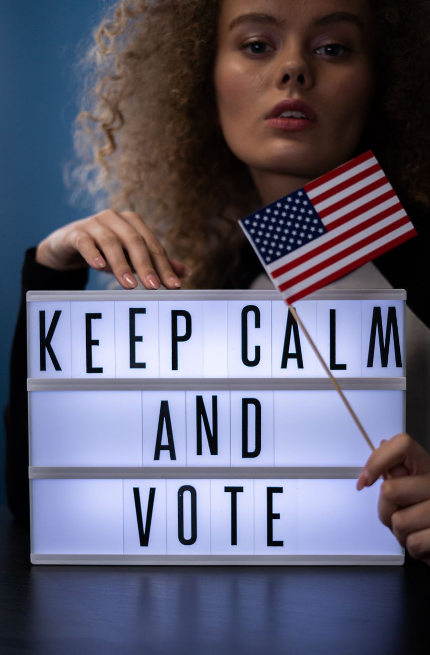 Stock photograph showing a person holding a small American flag next to an illuminated cinema-style light box that reads "KEEP CALM AND VOTE" from Pexels.com