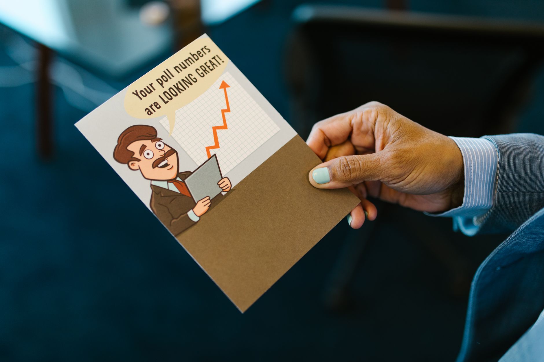 Stock photo of a hand holding a humorous greeting card with a cartoon illustration of a man and an upward-trending chart, captioned "Your poll numbers are LOOKING GREAT! from Pexels.com